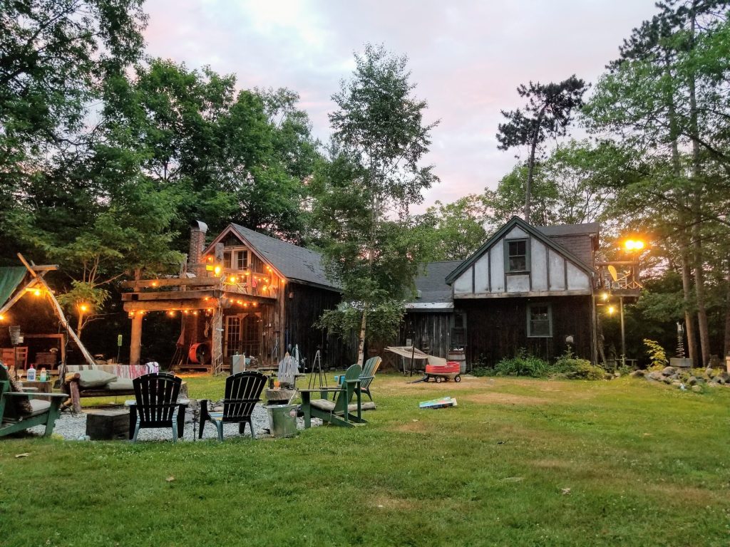 A cabin lit up at dusk with a fire pit and lawn chairs in the yard