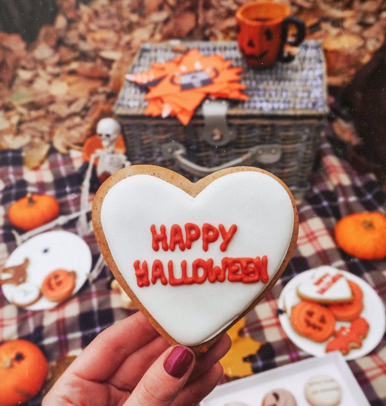 A hand holding a Happy Halloween heart cookie over an autumn picnic with pumpkins