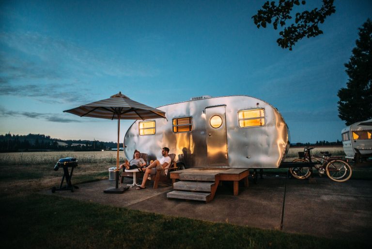 A vintage camper trailer lit up at dusk with people relaxing outside