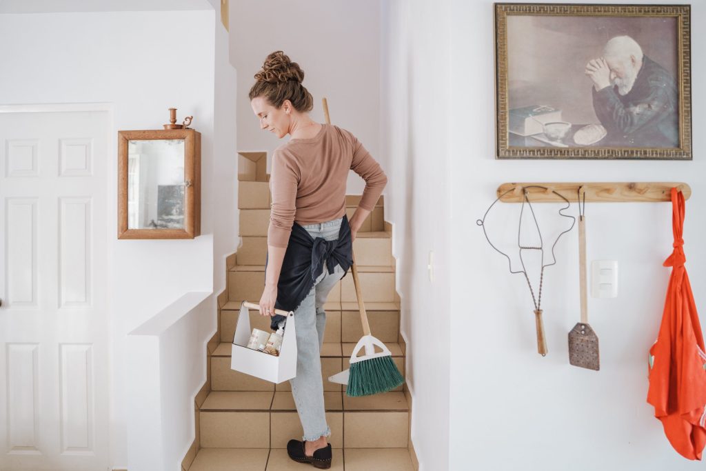 A woman in an apron carrying a cleaning caddy down a home staircase